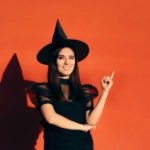 a woman in a witch costume and hat posing winking and posing in front of an orange background