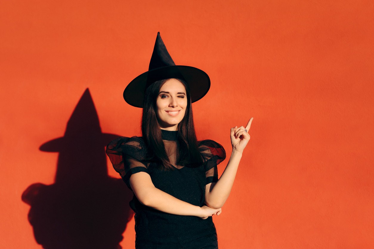 a woman in a witch costume and hat posing winking and posing in front of an orange background