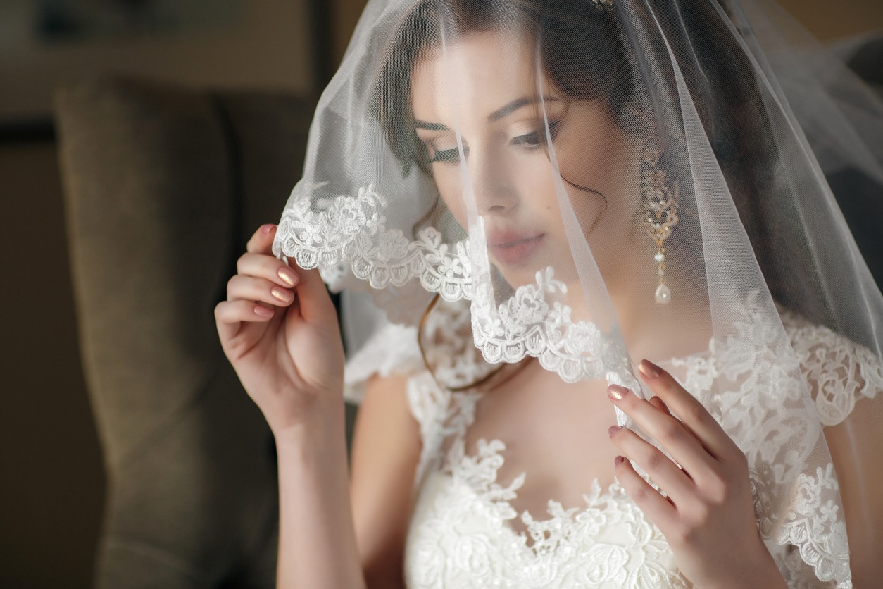 The young beautiful woman, the brunette with a wedding hair dress and professional make-up, in ears expensive earrings, is dressed in a white wedding dress and a transparent white veil, waits for the groom in a hotel room