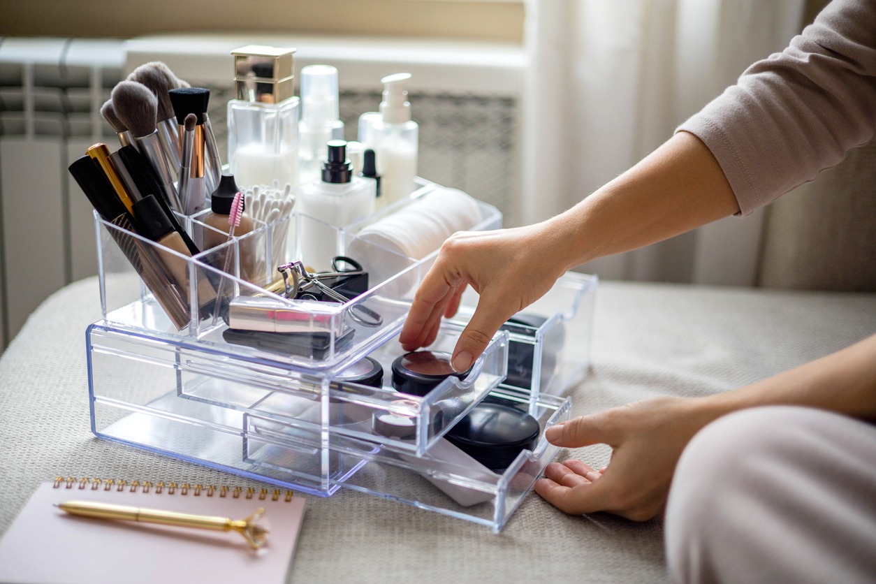 Closeup female hands putting luxury cosmetic into acrylic box with drawer storage organization. Beauty products tidying up neatly placement at minimalist container. Powder, foundation, lipstick, blush