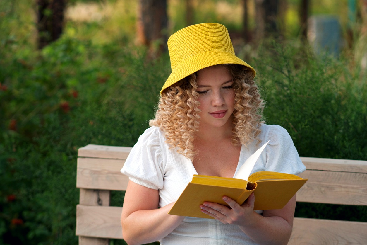 Romantic curly-haired girl reads the novel in paper book with yellow cover on eco-bench in the park. A young woman wears a yellow Panama bucket hat and a white summer outfit. Female student studying summer vacation