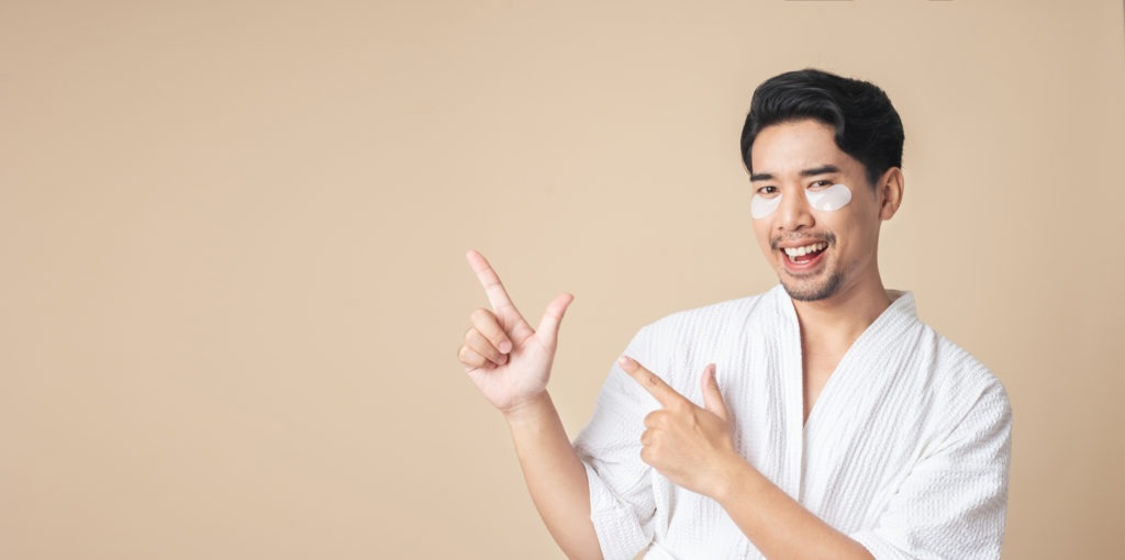 Isolated portrait Asian man in bathrobe smiling with patches mask under eyes and finger pointing to empty space on brown color background. Facial face care for man.