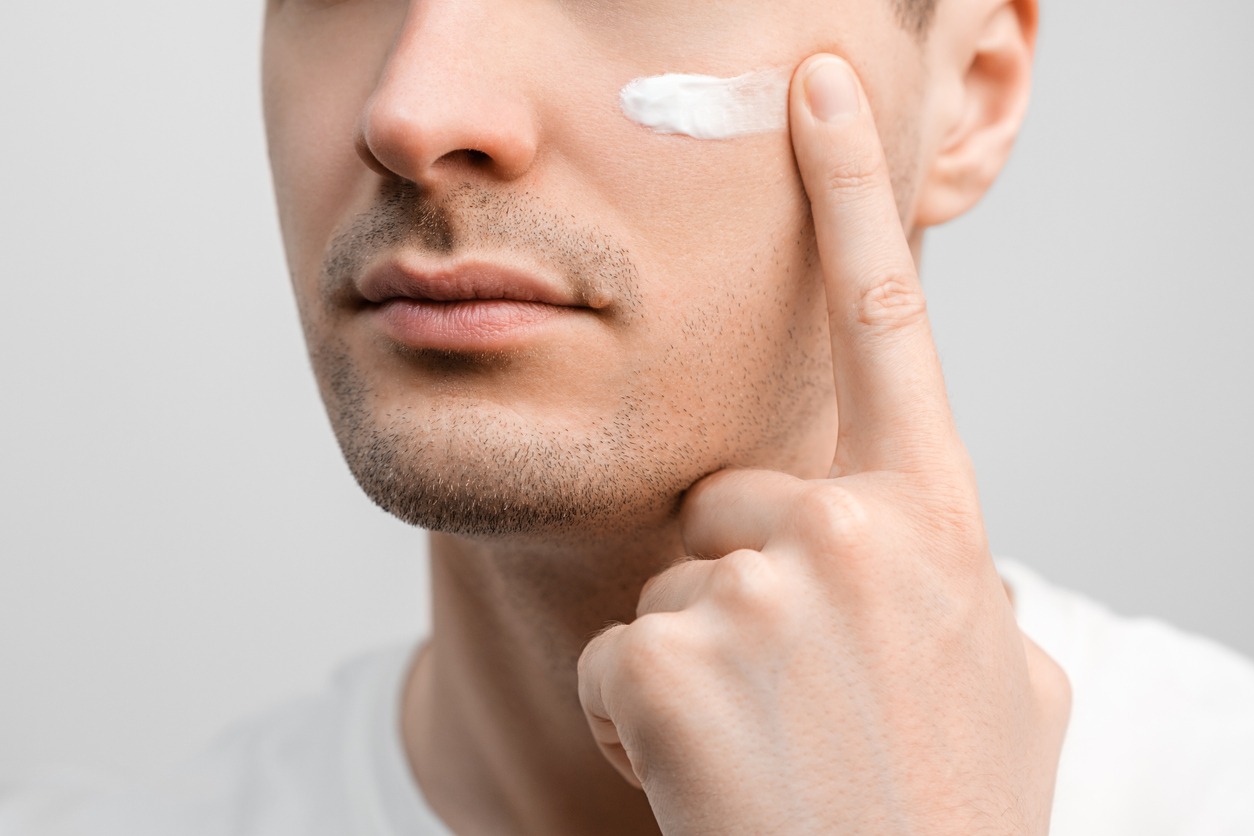 Handsome man with a stubble in a white t-shirt, sample of a face cream on his cheek, smudging with finger.