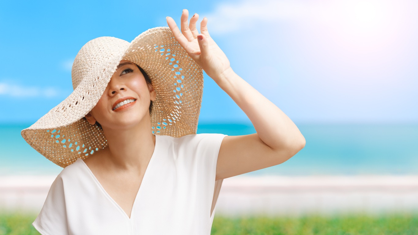 Beautiful short hair Asian woman with a white top, and straw hat looking up, smiling and raising a hand to block sunlight at the beach with blue sea and sky
