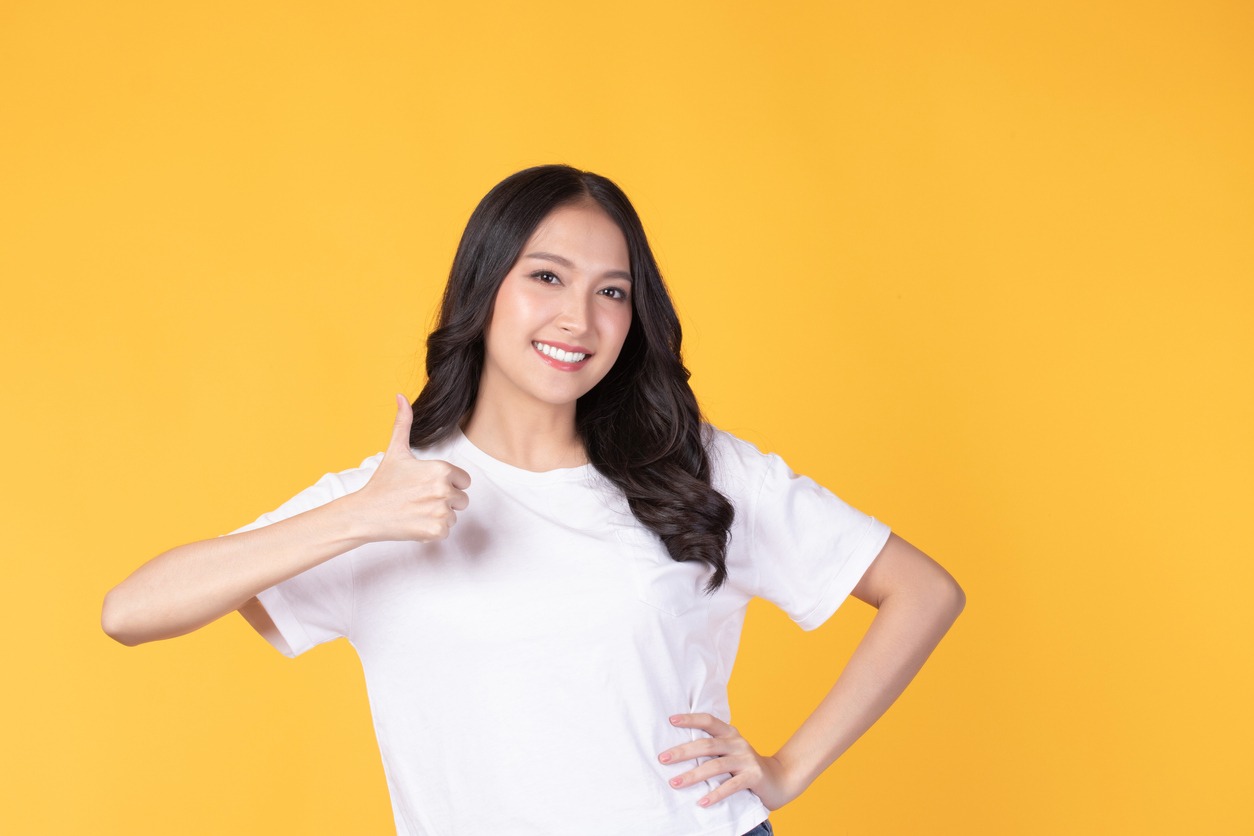 Happy young Asian woman wearing white shirt smile and showing thumbs up isolated on yellow background.