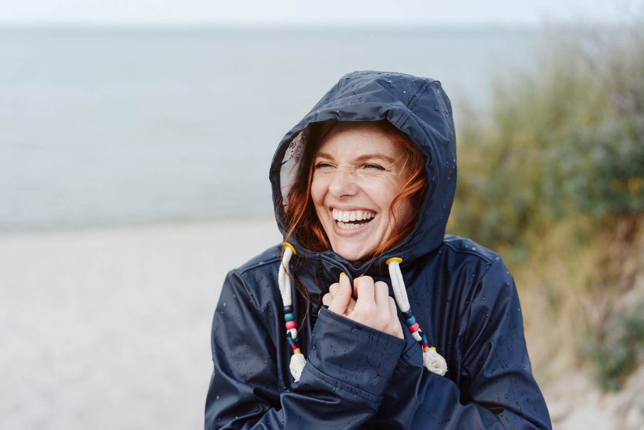 Laughing happy young woman embracing the cold autumn weather snuggling into her warm anorak with a beaming vivacious smile as she strolls along a beach