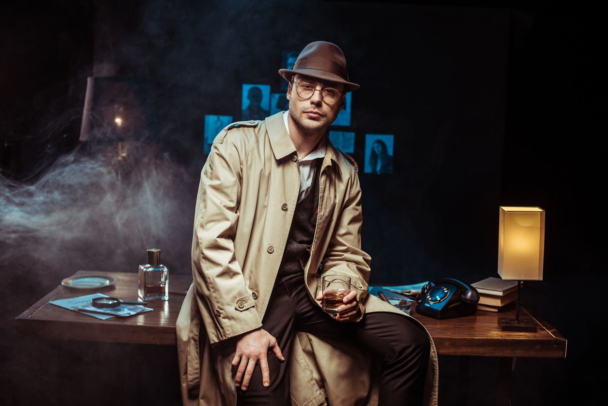 Detective in trench coat and hat sitting on table and holding glass of cognac