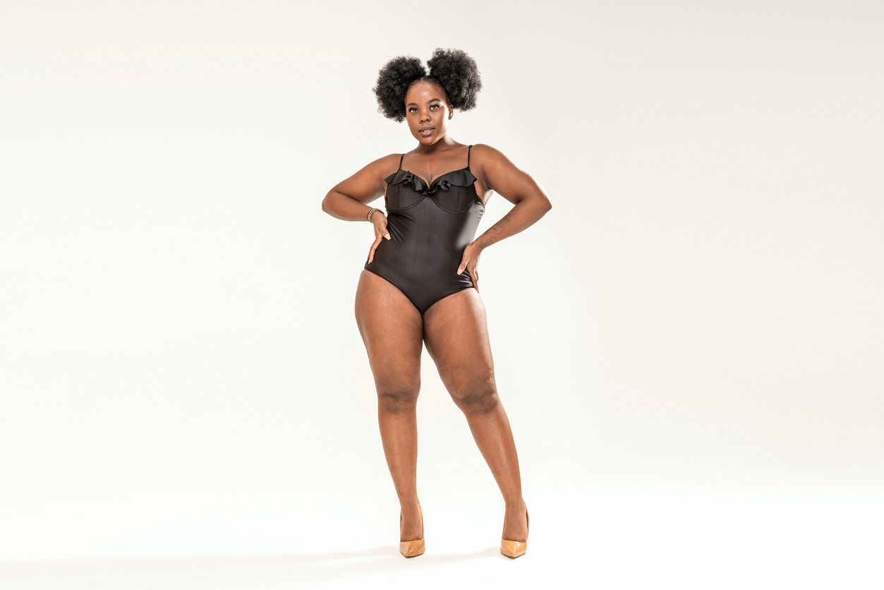 Young african plus size woman posing in black one piece swimwear, standing over light grey studio background